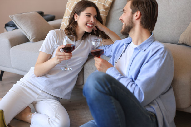 幸福的夫妻坐在客厅的地板上，放松地喝着红酒。微笑年轻的丈夫和妻子休息在家里享受浪漫的约会在家庭周末一起图片下载