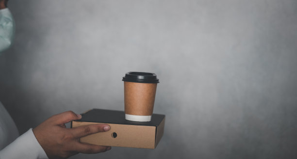 在冠状病毒大流行期间，带着医用口罩、披萨盒和一杯热咖啡的快递员随时准备在线送餐，提供快速送货上门和免费送货服务图片下载