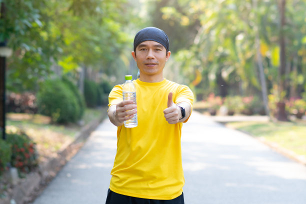 运动员在公园里喝水瓶。男性跑步者在公园跑步后出汗和口渴，并竖起大拇指图片下载