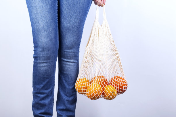 穿着蓝色牛仔裤的年轻女子手持有各种柑橘灰色背景的网袋。概念零浪费，环保。本空间图片下载
