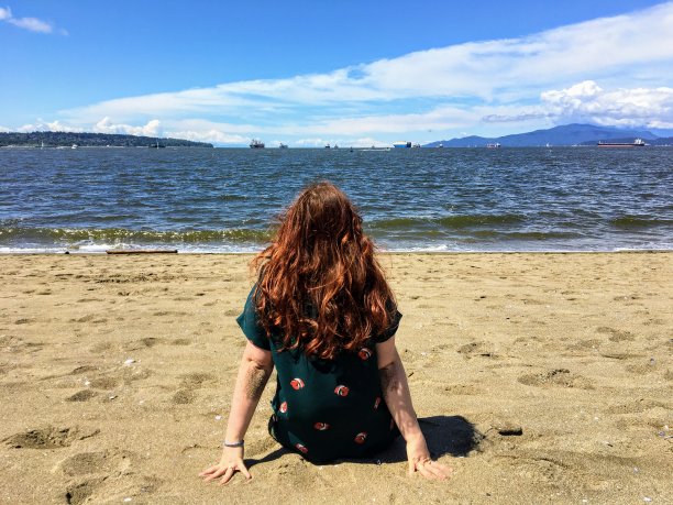 在一个阳光明媚的日子里，一位美丽的年轻女子坐在海滩上，沉思着凝视着加拿大不列颠哥伦比亚省温哥华市的大海和群山。图片下载