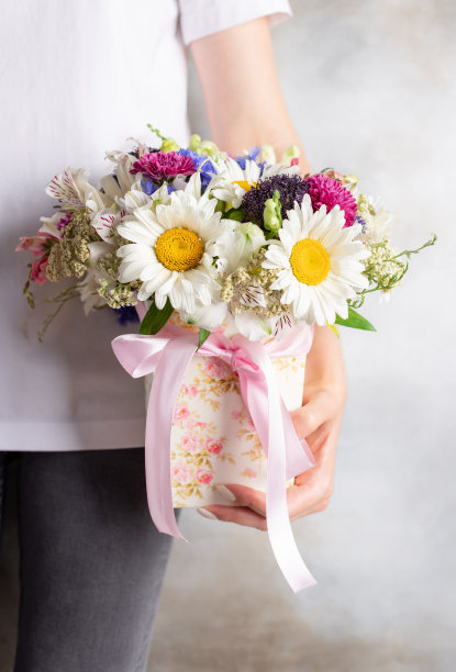 一名穿着白色t恤的年轻女子，手里拿着一束装在盒子里的花。白色雏菊，黄花，菊花。图片下载