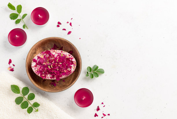 手工制作的肥皂，玫瑰花瓣放在一个木制的肥皂盘里，蜡烛，绿叶和一条浴巾在白色的背景上。有机沐浴产品图片下载