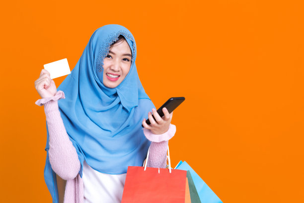 照片中的伊斯兰妇女戴着头巾戴着面具东方化妆使用智能手机和信用卡在橙色孤立的背景图片下载