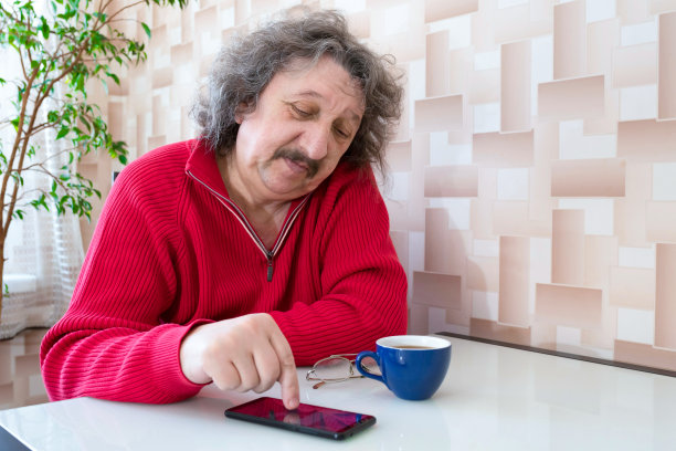 一位六七十岁的老人正坐在桌边，手里拿着一杯咖啡和一个电话。图片下载