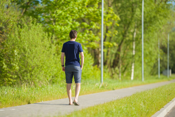 一个穿着短裤和t恤的年轻人慢慢地走在镇绿公园的长人行道上。温暖，阳光明媚的春日。花时间独处在大自然中。和平的气氛。后视图。图片下载