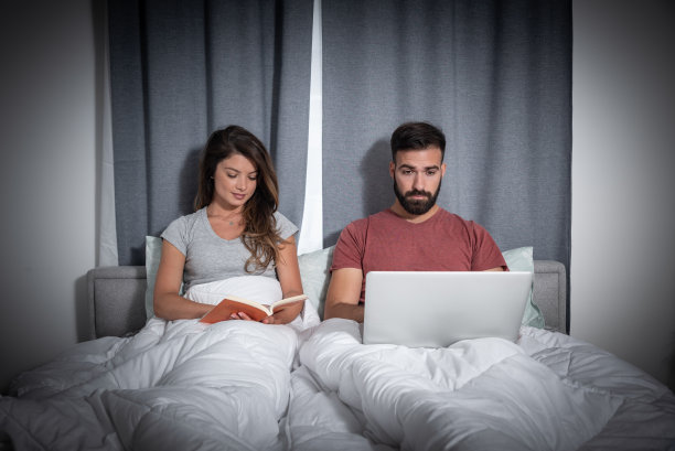 年轻夫妇和女人躺在床上，她在读一本书，他在他的笔记本电脑上完成他的自由职业工作前睡觉图片下载