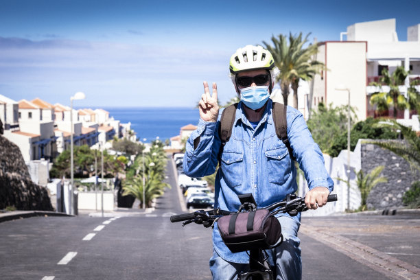 老年男人戴着黄色头盔在沙漠公路上骑自行车，疫情尚未结束，面部戴口罩保持社交距离图片下载