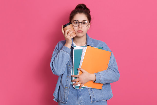 漂亮的女学生用带走的咖啡抚摸着自己的脸颊，拿着文件夹，穿着夹克和眼镜的女士，带着悲伤或严肃的表情看着相机。图片下载