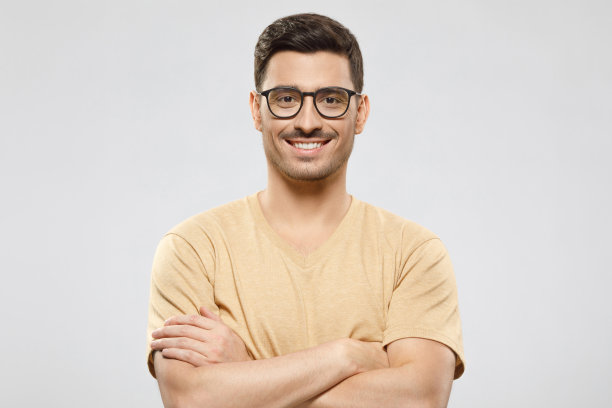 这个年轻人身穿米色t恤，戴着眼镜，双臂交叉站在那里，笑容灿烂，自信而专业，在灰色的背景下显得很孤立图片下载
