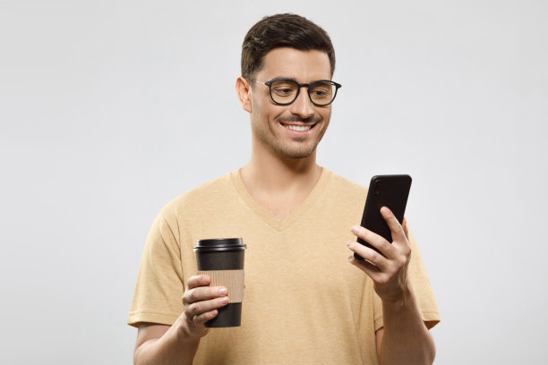 一个穿着t恤、戴着眼镜、面带微笑的年轻人，拿着外卖咖啡杯，在灰色背景中查看智能手机上的新信息图片下载