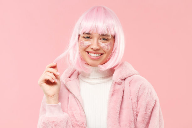 年轻美丽的女性的肖像戴假发，面颊上的闪光和蓬松的外套，微笑和触摸的头发，孤立在粉红色的背景图片下载