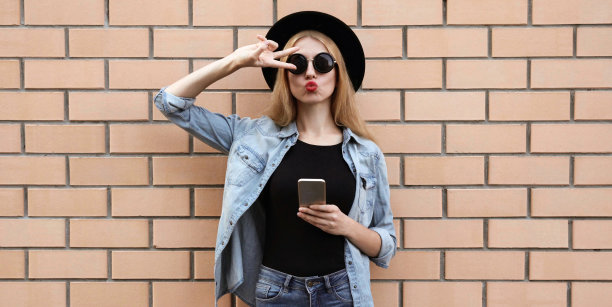 肖像时尚的年轻女子模特用手机吹红色的嘴唇发送甜蜜的飞吻戴着黑色圆帽，牛仔裤夹克在砖墙的背景图片下载
