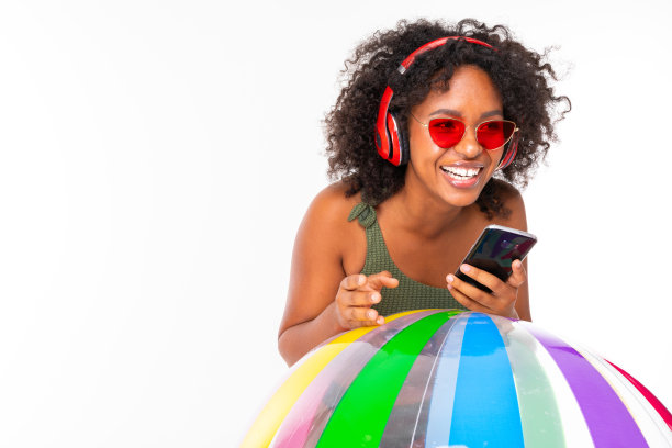 漂亮的非洲女性穿着泳衣站在大彩色橡胶球，听音乐与耳机，做孤立的白色背景自拍图片下载