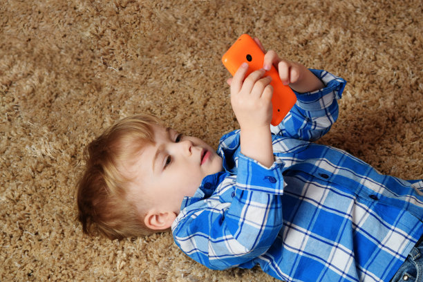 一个小男孩正躺在地毯上玩电话。小男孩在用智能手机。图片下载