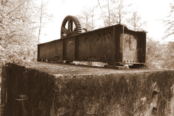 这是一张深褐色的照片，上面是一座金属桥的残骸，顶部有一个生锈的大齿轮，下面是一个巨大的金属矩形，它矗立在波兰茂密的森林中间的混凝土地基上图片下载