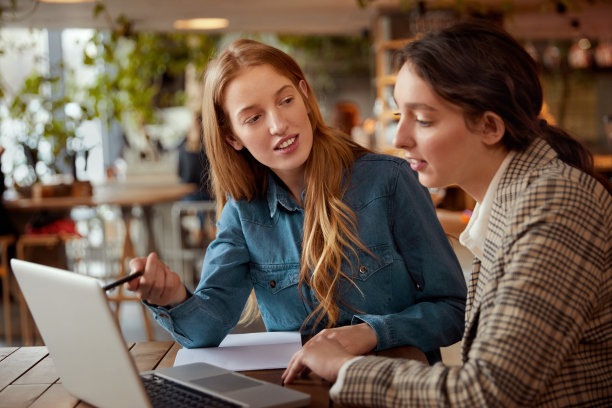 学生。用笔记本电脑学习的女孩。美丽的年轻女人在咖啡馆上课。微笑的金发和黑发看着屏幕。图片下载