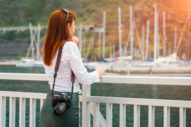 一个漂亮的读书女孩拿着相机看着阳光下的码头。女人准备拍摄旅游照片在加那利群岛。游艇在软焦点背景。图片下载