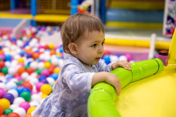 一个可爱的蹒跚学步的小女孩穿着裙子，在一个有球的游泳池的背景上窥视着一边图片下载