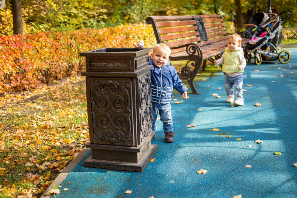 一个可爱的小婴儿穿着蓝色的衣服站在一个公园的街边瓮旁，在那里扔垃圾，微笑着看着镜头。另一个孩子在后面。软焦点，背景模糊。图片下载