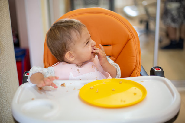 可爱的婴儿坐在橙色的婴儿椅和一张桌子，手和脸都很脏，看着别处，笑着。桌上有面包屑和一个橙色的盘子。特写，前视图，软焦点，模糊背景素材图片
