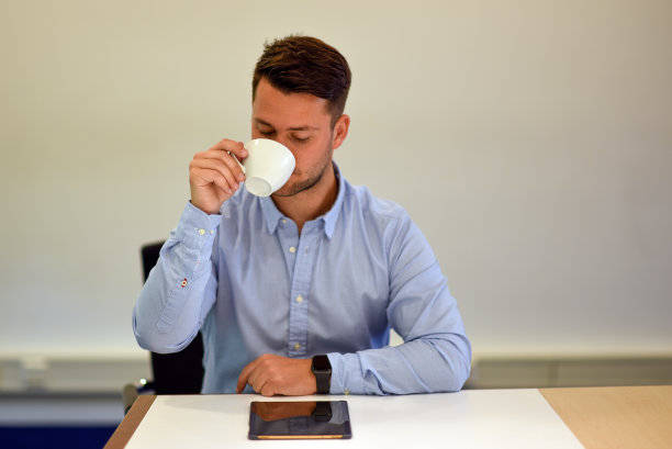 一位英俊的商人一边从杯子里喝水一边看着平板电脑。图片下载