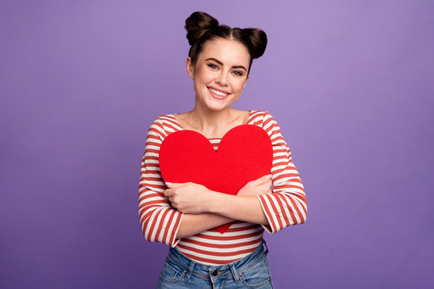 漂亮可爱的少女的照片抱着大纸心接近胸部庆祝情人节创意明信片穿白色红色休闲条纹衬衫孤立柔和的紫色背景图片下载