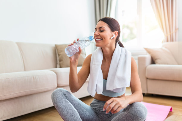年轻的成年女子喝着塑料瓶里的水，坐在健身垫上，在家训练后休息图片下载