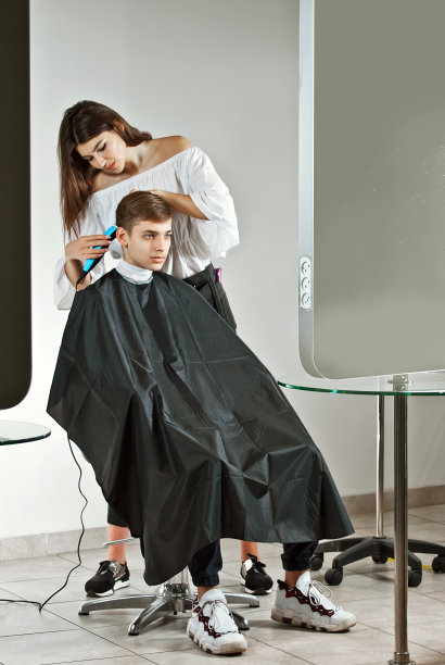 理发师用理发器给那家伙理发。头靠近。在模型附近放置文本。布局设计，复制空间。穿着黑色斗篷的顾客被孤立在白色的背景下。图片下载