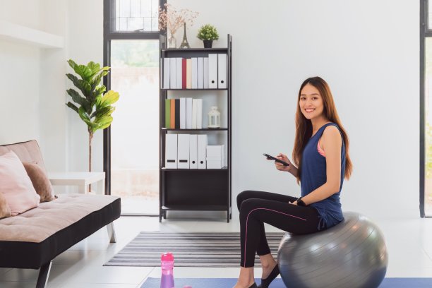 美丽的亚洲健身女人坐在运动球和使用智能手机前，在客厅背景。封锁、隔离和社交距离的新正常生活方式概念。图片下载