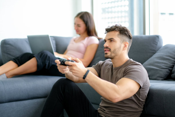 英俊的男人拿着游戏操纵杆和享受玩游戏，而他的女朋友或妻子工作与笔记本电脑。新型冠状病毒在城市流行期间的工作理念和居家娱乐活动图片下载