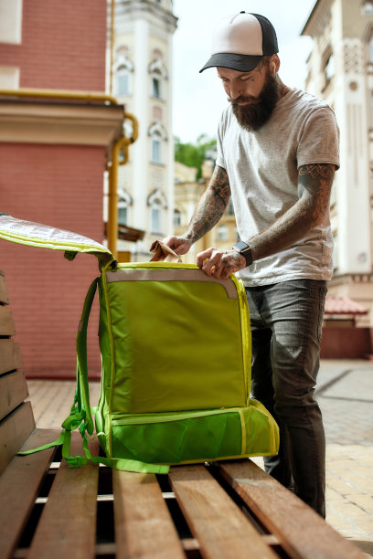 秩序和享受。野蛮的大胡子送货员，戴着帽子打开保温袋或背包，把食物拿出来或放进去，站在户外长凳附近。快递、派送的服务理念图片下载