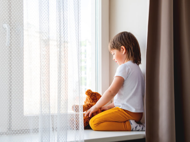 蹒跚学步的小男孩和泰迪熊坐在一起。拿着毛绒玩具的孩子透过窗户往外看。棕熊和孩子因新冠肺炎在家隔离等人。图片下载