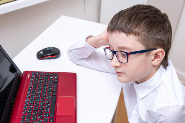 一个聪明的男孩，一个穿着白色正装衬衫和眼镜的学生坐在桌子旁，书桌上有笔记本电脑，在家里做作业，在线学习，远程课程和在线教育的概念图片下载