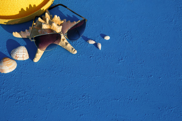 以海洋主题的沙滩饰品为框架:黄色草帽、太阳镜、海星和贝壳在蓝色背景上。图片下载