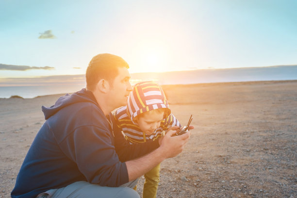 夏日日落时分，父子俩在海边操控无人机。自由，家庭，旅行，旅行，团聚。图片下载