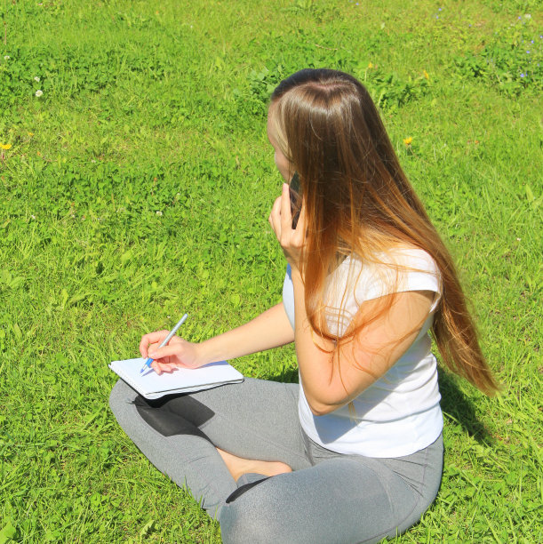 一个漂亮的年轻白人女孩穿着白色的t恤，长头发坐在绿色的草地上，在草坪上，打电话，用钢笔在笔记本上写图片下载
