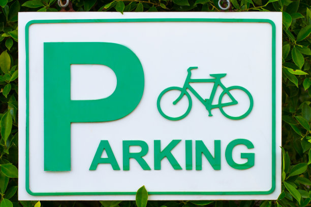 绿色的停车标志，白色的背景，还有一个自行车标志和一个标志挂在绿色的树叶丛旁边。自行车停车标志。图片下载