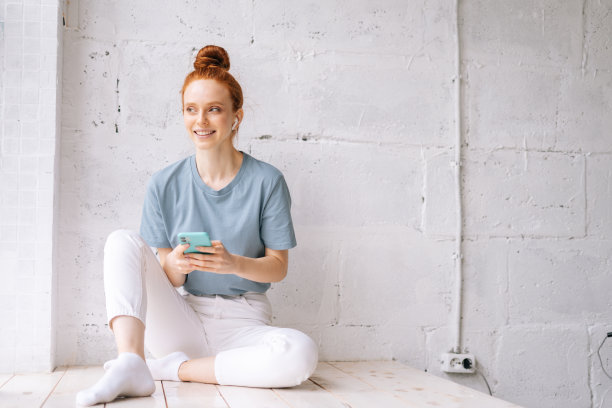 快乐的红发年轻女子戴着无线耳机听轻松的音乐使用手机。图片下载