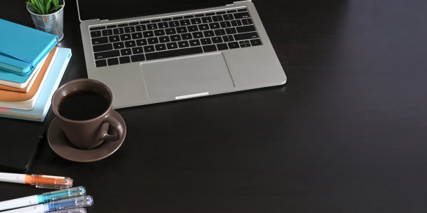 电脑笔记本放在黑色的办公桌上，周围是陶瓷咖啡杯，一堆笔记本和笔。有序的工作空间的概念。图片下载