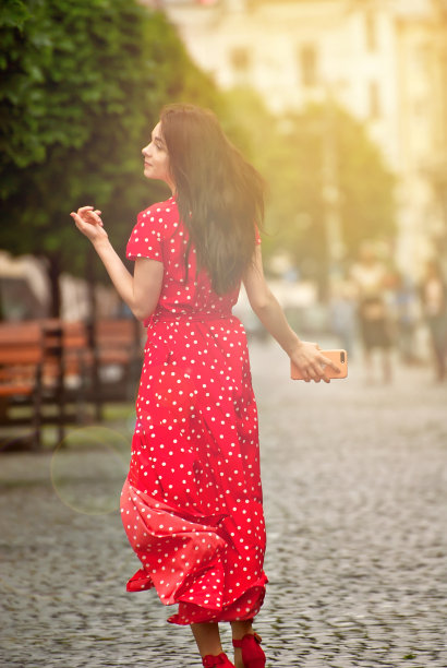 快乐的女孩用鹅卵石走在街上。穿着有白点的红裙子的女人。衣服和头发被风吹走了。模糊的城市街道上的女孩。图片下载