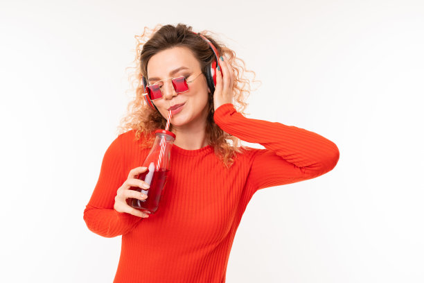 可爱的卷发金发女孩戴着时髦的太阳镜，穿着红色的裙子，喜欢在白色的背景下听音乐图片下载
