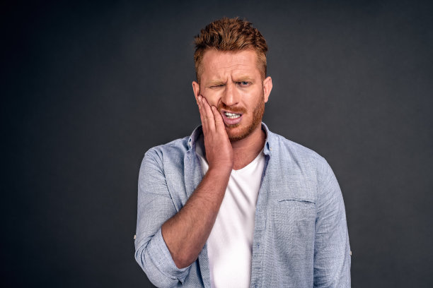 照片的高加索红发男子遭受痛苦的牙痛，有腐烂的牙齿，需要去看牙医，保持眼睛闭着从痛苦，穿着休闲衬衫，孤立在灰色的背景，触摸脸颊图片下载