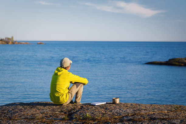 一个快乐的男人坐在海边，身边放着一本书和一个钛杯子。图片下载
