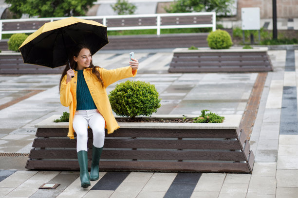 一个年轻漂亮的少女走在城市的雨天。穿着彩色衣服和胶靴的女孩用手机拍自己的照片图片下载