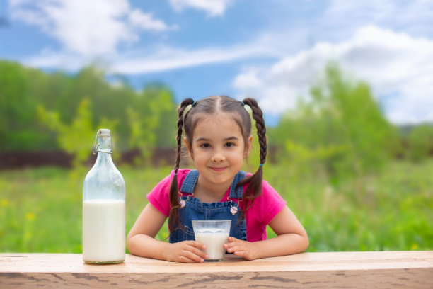 梳着两条辫子的漂亮小女孩，坐在一张桌子旁，端着一杯牛奶，微笑着。图片下载