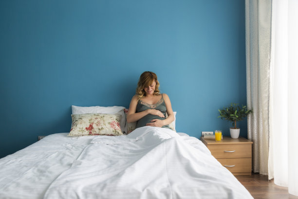 一个微笑的孕妇在床上半坐的姿势，她的手在她的肚子。图片下载