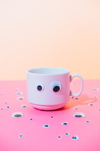 一杯热咖啡或茶与卡通玩具眼睛在粉红色和黄色柔和的超现实主义背景。图片下载
