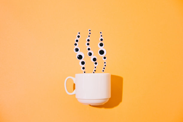 一个白色的咖啡杯冒着热气，在黄色蜡笔超现实主义的背景上形成了卡通玩具眼睛的形状。图片下载
