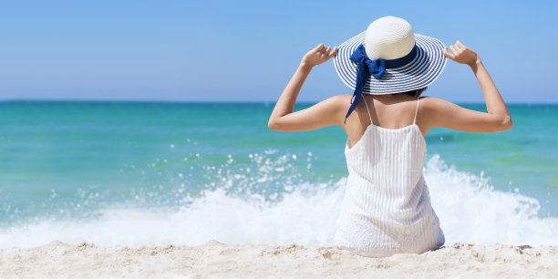 在海滩度假的夏季女性：快乐的女人戴着夏天的裙子和草帽坐在海滩上看着大海。在热带岛屿海滩上拍摄的夏季生活方式户外照片，放松身心。素材图片